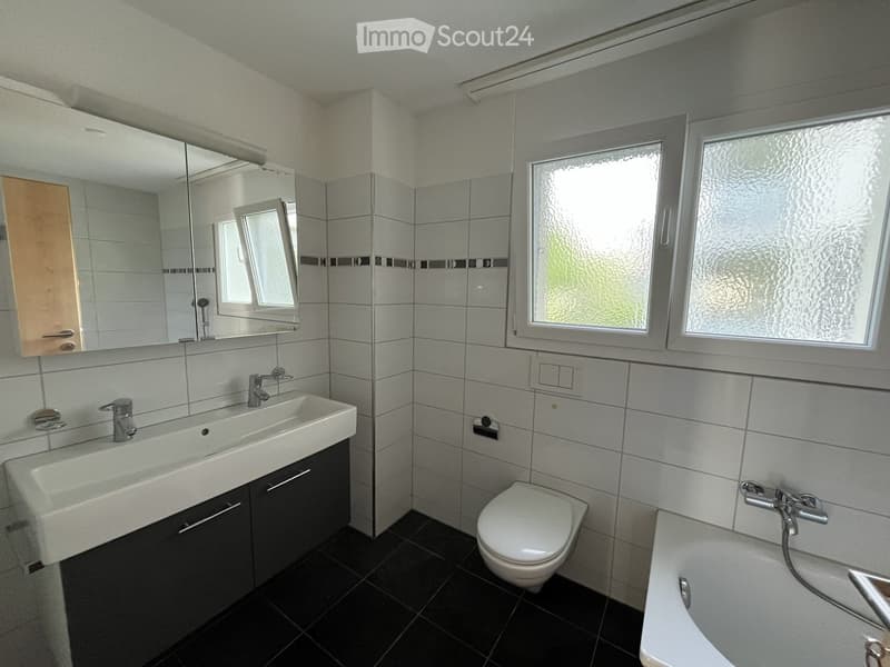 Modern bathroom with white tiles, black floor, large mirror, double sink, toilet, bathtub, and frosted windows.