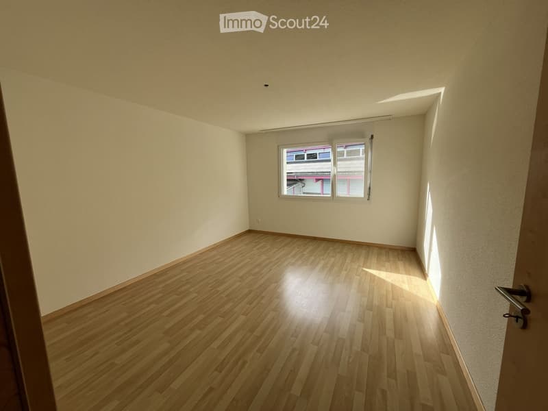 Empty room with wooden flooring and white walls, featuring a window with blinds and a door handle visible on the right side.