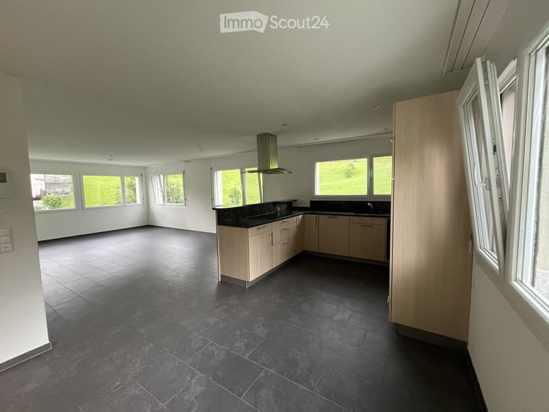 Empty kitchen with light wooden cabinets, black countertops, and large windows overlooking a green landscape.