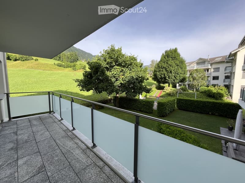 Balcony with modern glass railing, tiled floor, overlooking a green lawn and trees.
