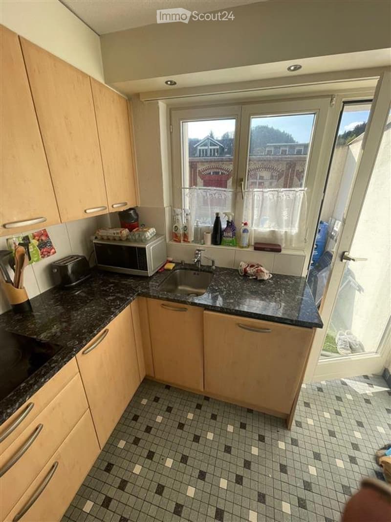 Compact kitchen with light wooden cabinets, granite countertop, microwave, toaster, and sink. The space features tiled flooring and a window with a view of the outside.