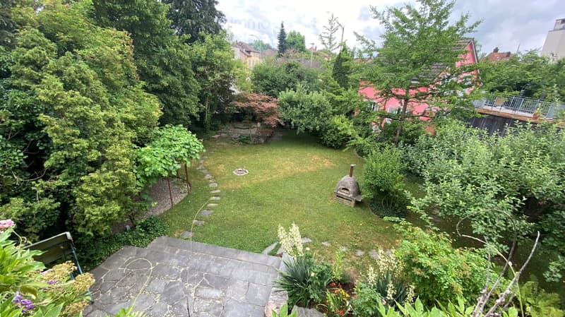 A garden with a grassy lawn, a stone pathway, a stone bench, a stone oven, and a variety of plants and trees.