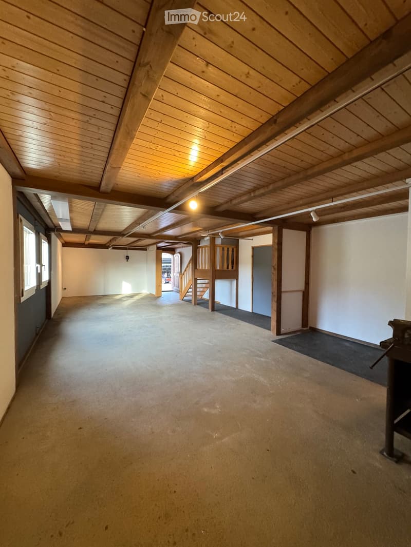 Empty room with wooden ceiling beams, concrete floor, and white walls. Features a staircase leading to an upper level and a small window on the left side.