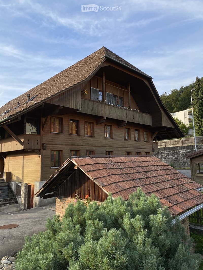 Two-story wooden house with a steep, tiled roof and multiple windows. Features a balcony on the upper level and a small attached structure with a separate roof.