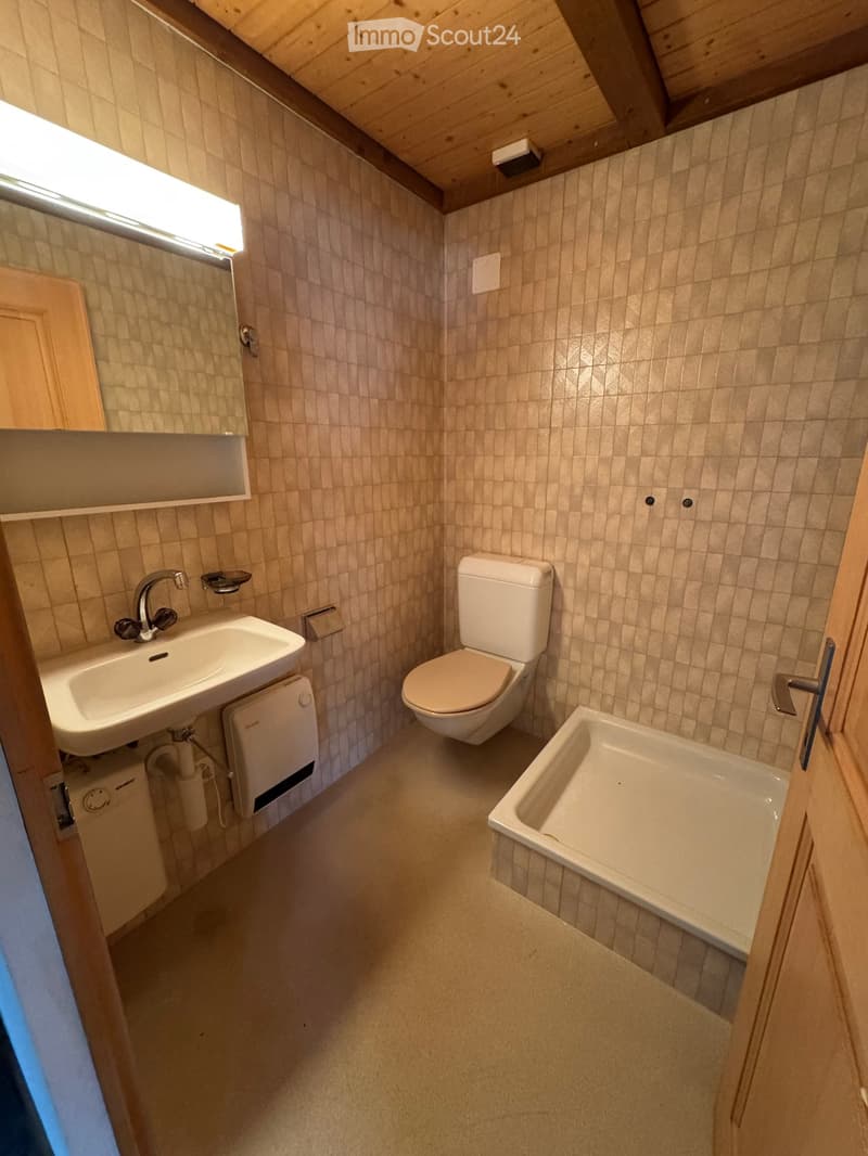 Compact bathroom with beige tiled walls, a white sink, a toilet, and a bathtub, featuring wooden cabinetry and a built-in shelf above the sink.