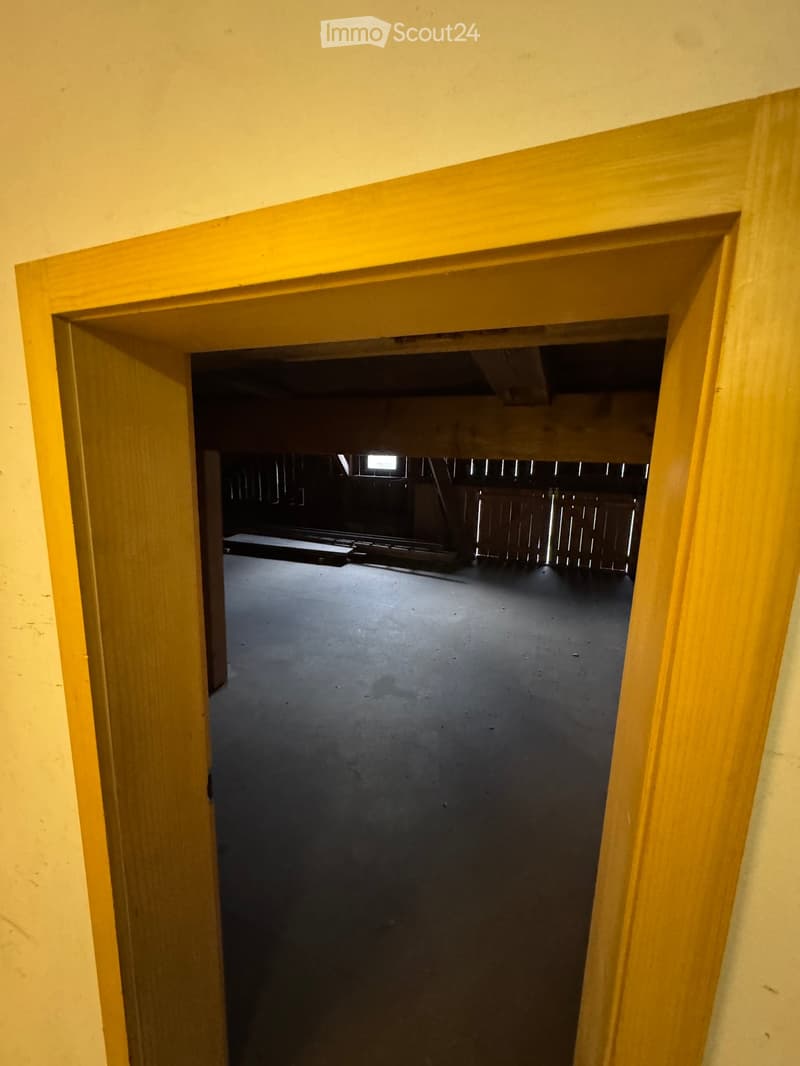 An empty, unfurnished room with a concrete floor and wooden beams visible in the background. The doorway is framed in light wood.