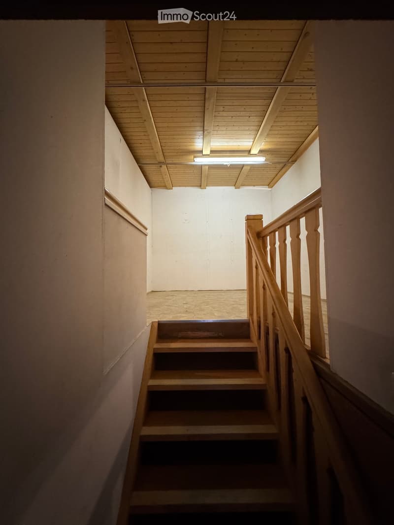 Wooden staircase with a handrail, white walls, and a wooden ceiling with exposed beams and a single light fixture.