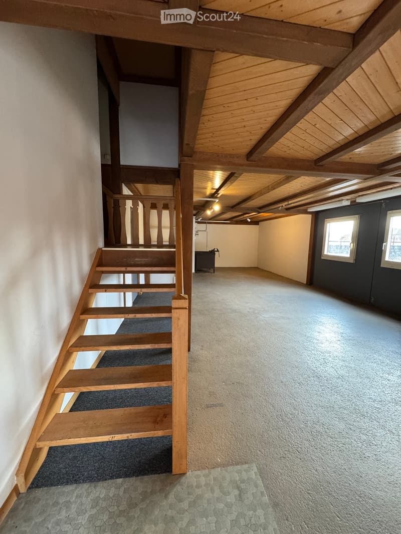 Wooden staircase leading to an open loft area with exposed wooden beams and a skylight, featuring a mix of carpet and concrete flooring.