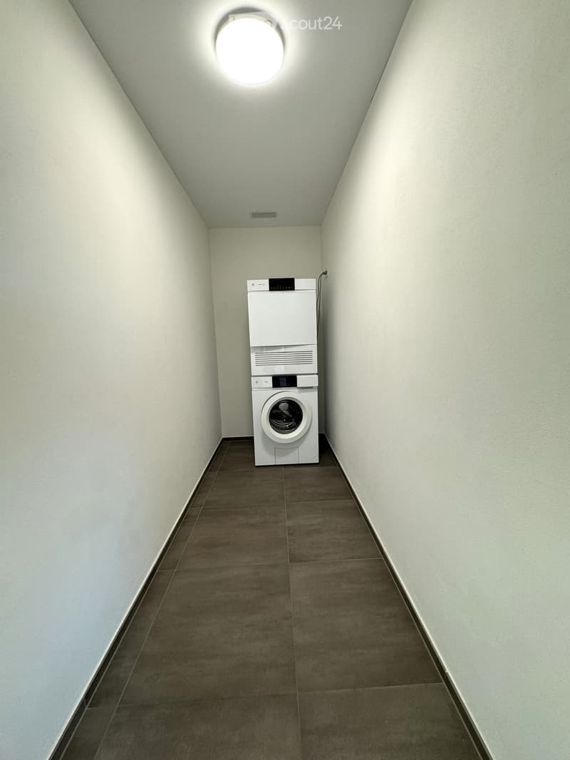 A narrow hallway with light grey walls and a brown tiled floor, featuring a white washing machine and dryer unit positioned against one wall.