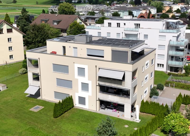 A modern, multi-story house with solar panels on the roof, large windows, balconies, and a well-maintained garden with trees and shrubs.