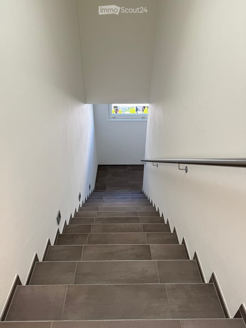 Modern staircase with gray tiled steps and white walls, featuring a metal handrail and a window at the top letting in natural light.