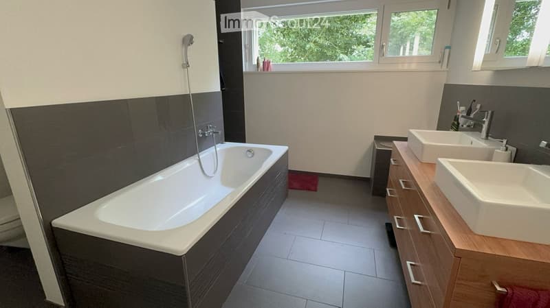 Modern bathroom with a bathtub, double sink, and wooden cabinet. Features large windows with a view of greenery.