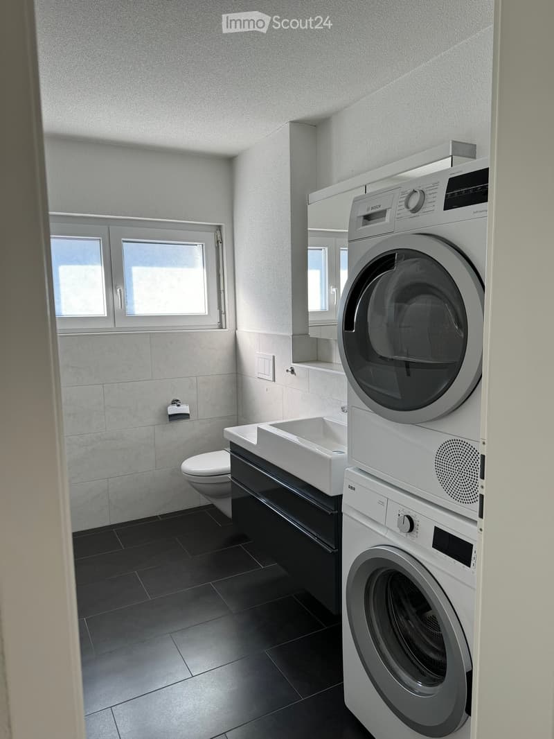 Modern bathroom with a toilet, sink, and stacked washing machine and dryer unit. The space features dark tiled flooring and light grey walls.
