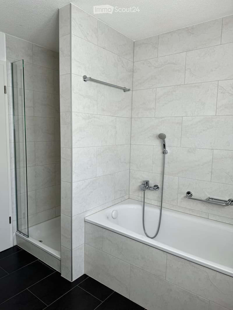 Modern bathroom with a white bathtub, marble tiles, glass shower enclosure, and chrome fixtures including a showerhead and grab bars.