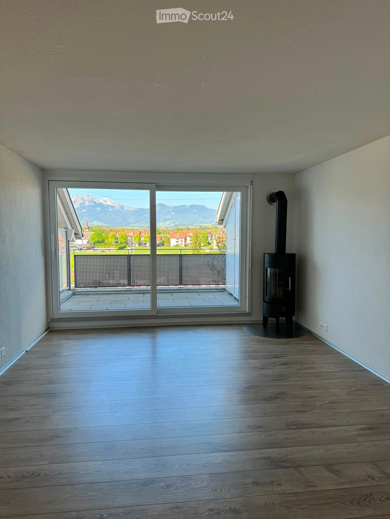 Empty room with wooden flooring, white walls, large sliding glass door leading to a balcony with a view of mountains and a town, and a black wood-burning stove on the right side.