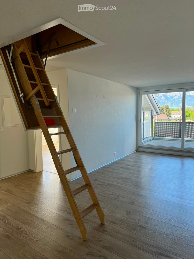 Empty room with wooden flooring, white walls, and a large window offering a view of mountains. A wooden ladder is positioned against the wall, leading to an attic space.