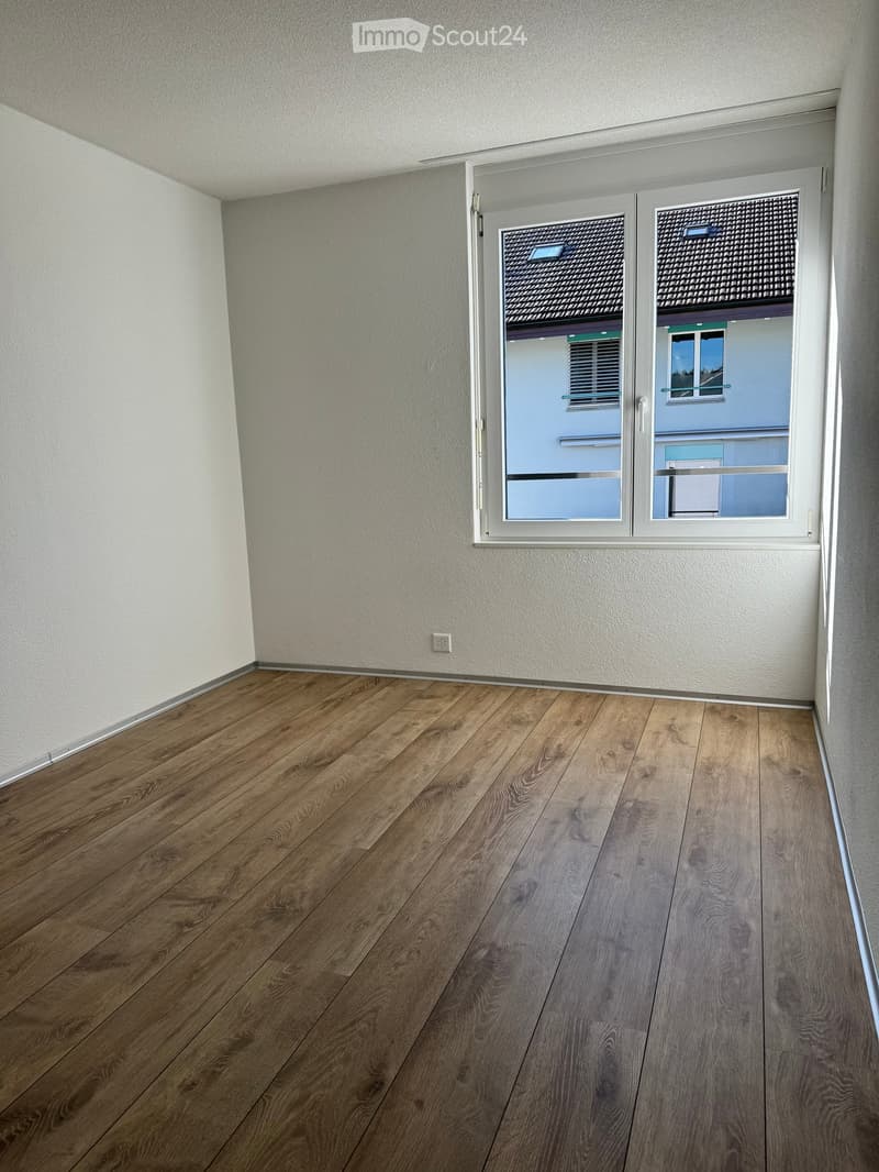 Empty room with wooden flooring and white walls, featuring a large window with a view of neighboring buildings.