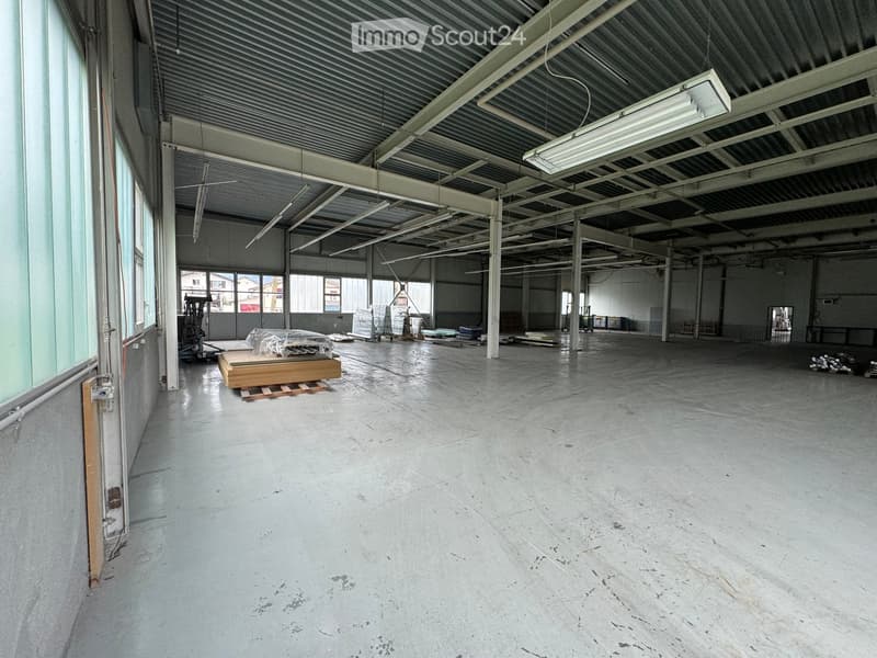 Empty industrial space with concrete floor, metal ceiling, windows, and a few stacks of material