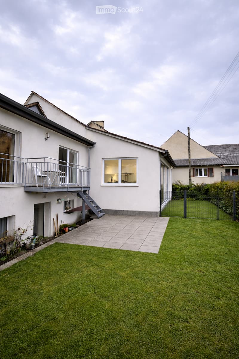 Lawn with a tiled pathway leading to a house with a balcony, stairs, and visible interior through windows.