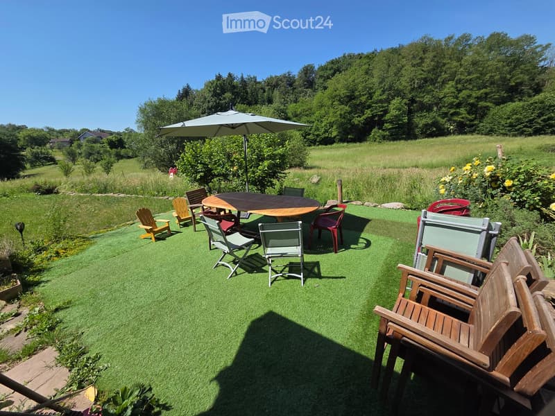 Well-maintained lawn with a round wooden table, multiple chairs, and an umbrella providing shade. The area is surrounded by greenery and trees, with a clear blue sky overhead.