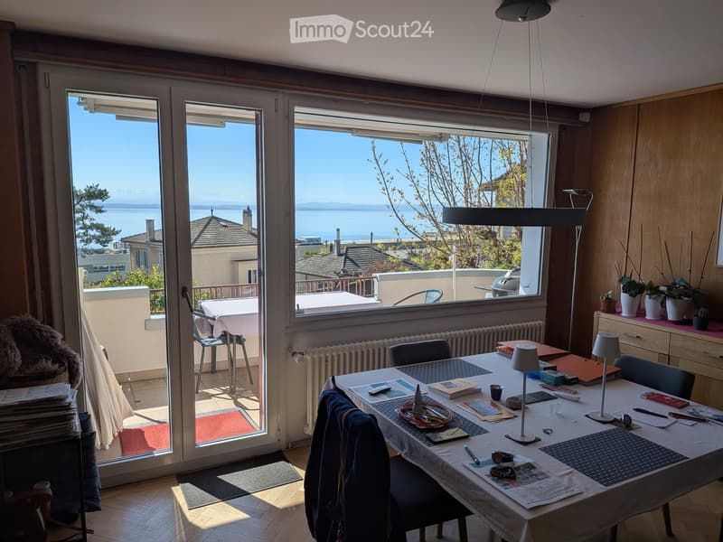 Bright room with large windows offering a scenic view, furnished with a dining table, chairs, a lamp, and decorative plants.