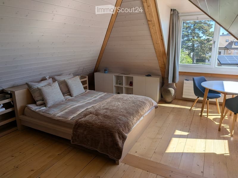 Cozy attic bedroom with wooden floors, slanted wooden ceiling, large window, bed with pillows and blanket, small table with chairs, cabinet, and decorative vase.