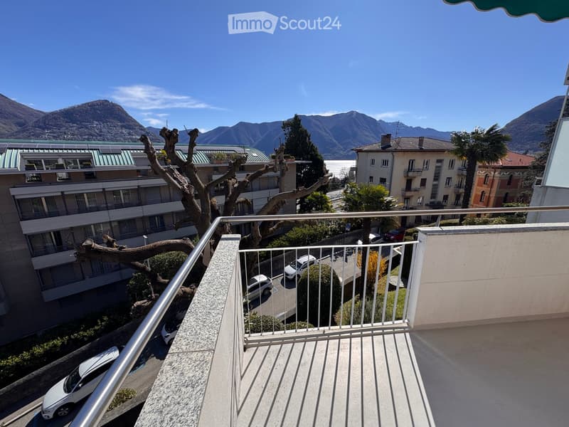 Balcony with metal railing overlooking a scenic view of mountains, a lake, and surrounding buildings. Several parked cars are visible below.