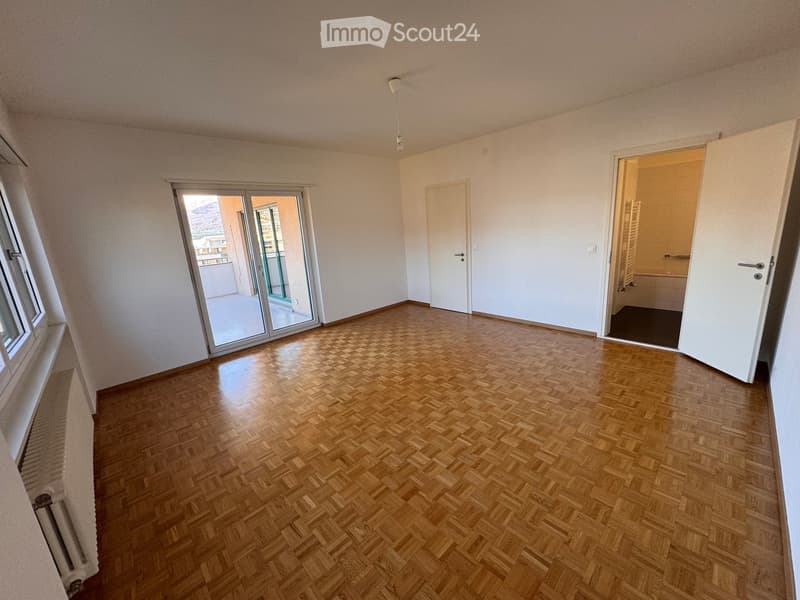 Empty room with wooden herringbone flooring, white walls, large window, sliding glass door leading to a balcony, and an open door to a bathroom.