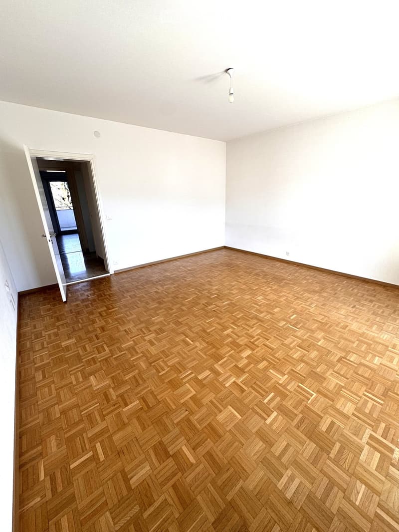 Empty room with wooden herringbone flooring, white walls, and a single hanging light fixture. An open doorway leads to another space.