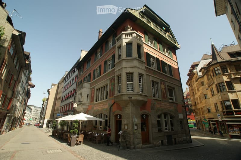 A multi-story building with ornate architecture, featuring a prominent corner tower and decorative facades. The ground floor has outdoor seating with tables and chairs under umbrellas, and the building has large windows and detailed stonework.