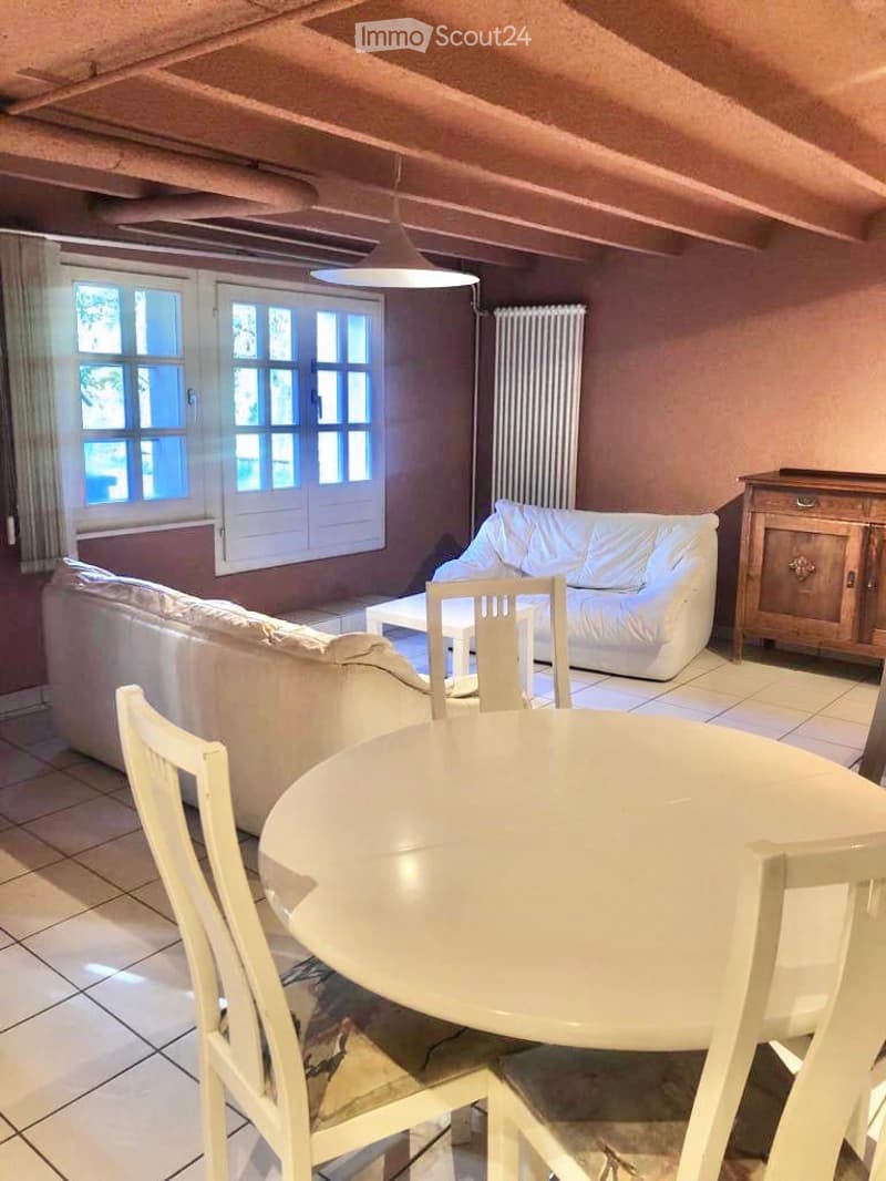 A cozy living room with a white sofa, a round dining table with chairs, a radiator, and a wooden cabinet. The room features large windows with white frames and a hanging light fixture.