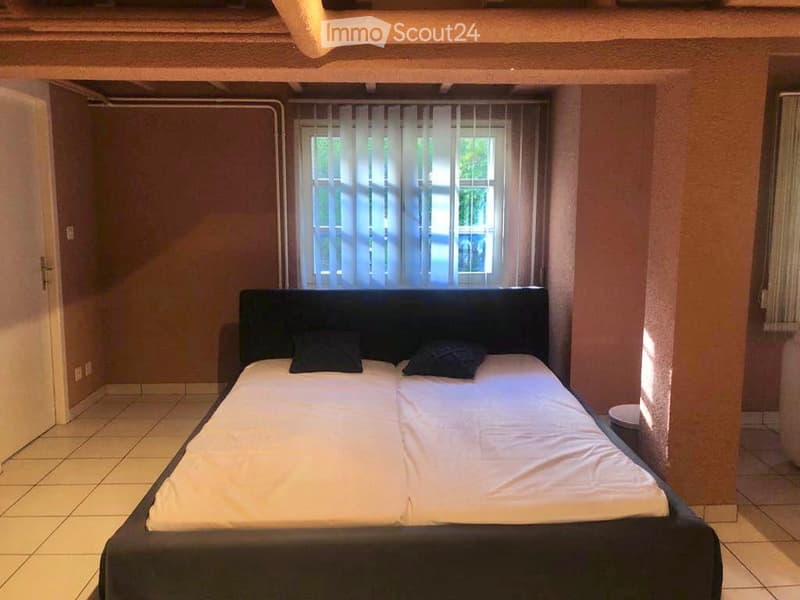 A bedroom with a double bed, two pillows, and a window with sheer curtains. The room has tiled flooring and warm-colored walls.