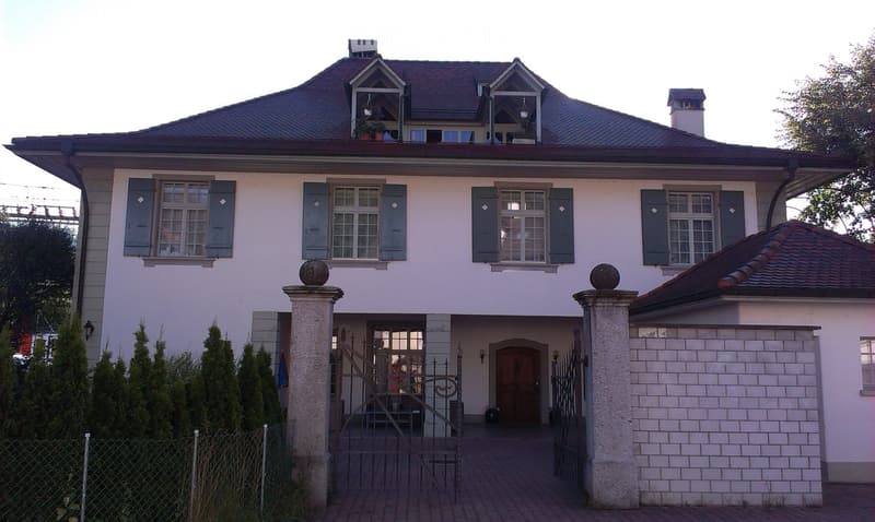 2 story house, white walls, brown roof, brown door, 2 gates, brick driveway, shrubs, fence