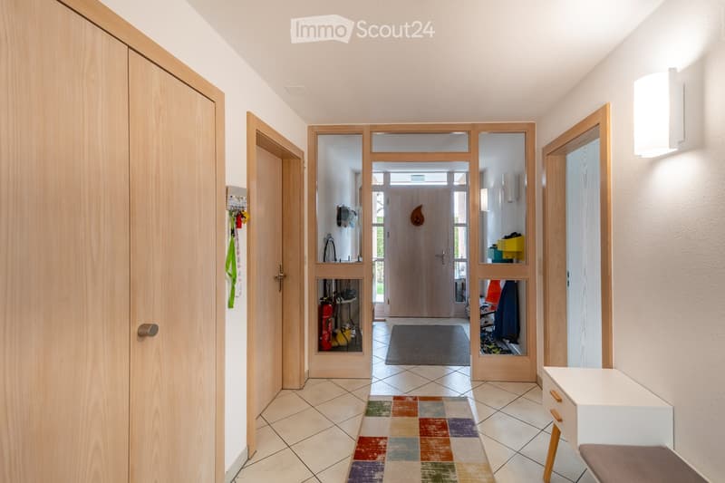 Bright hallway with tiled flooring, wooden doors, and a colorful rug leading to another room.