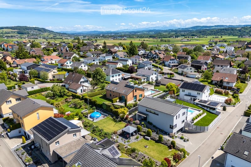 Aerial view of a suburban neighborhood with multiple houses, gardens, and solar panels on rooftops. The area features well-maintained lawns, driveways, and a scenic backdrop of mountains and fields.