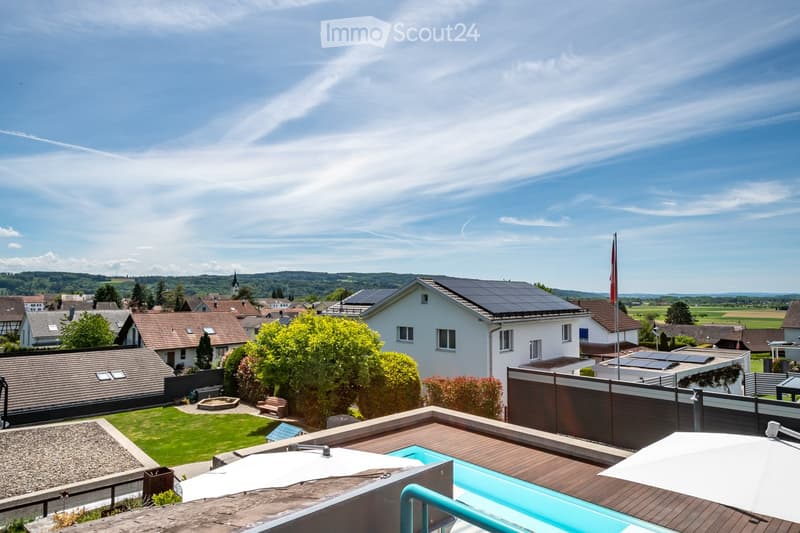 A sunny rooftop terrace with a swimming pool, wooden decking, and large umbrellas, overlooking a suburban neighborhood with houses, trees, and distant hills.