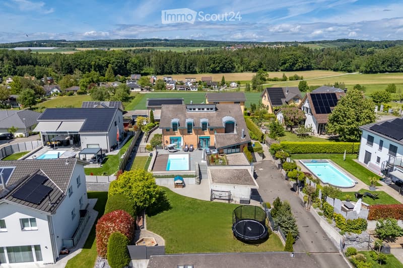 Aerial view of a suburban neighborhood with multiple houses, solar panels, swimming pools, and well-maintained gardens.