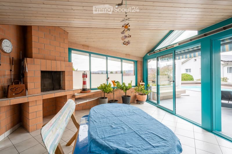 Bright living space with a fireplace, large windows, a covered table, and potted plants, leading to an outdoor area with a pool.