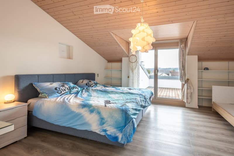 Cozy bedroom with wooden ceiling, large bed with blue and white bedding, nightstand with lamp, shelves, and a view through large windows.