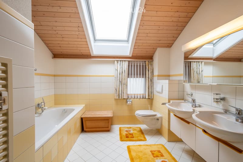 Bright bathroom with wooden ceiling, skylight windows, yellow and white tiles, bathtub, toilet, double sink, and mirrored cabinet.