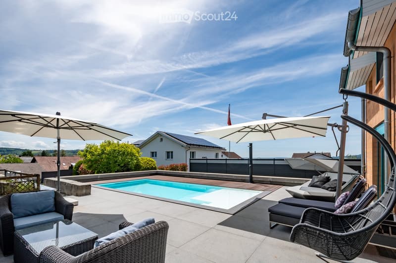 A modern outdoor area with a swimming pool, lounge chairs, wicker seating, and large umbrellas, overlooking a residential neighborhood.