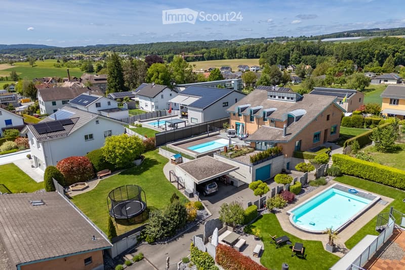 Aerial view of a residential area with multiple houses, gardens, swimming pools, and a trampoline in the foreground.