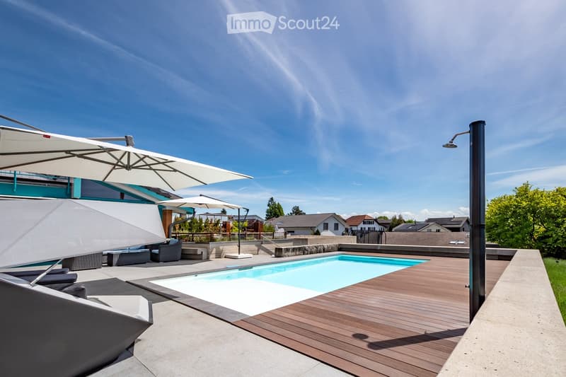 Modern outdoor area with a swimming pool, wooden decking, lounge chairs, umbrellas, and a shower, surrounded by greenery and neighboring houses.