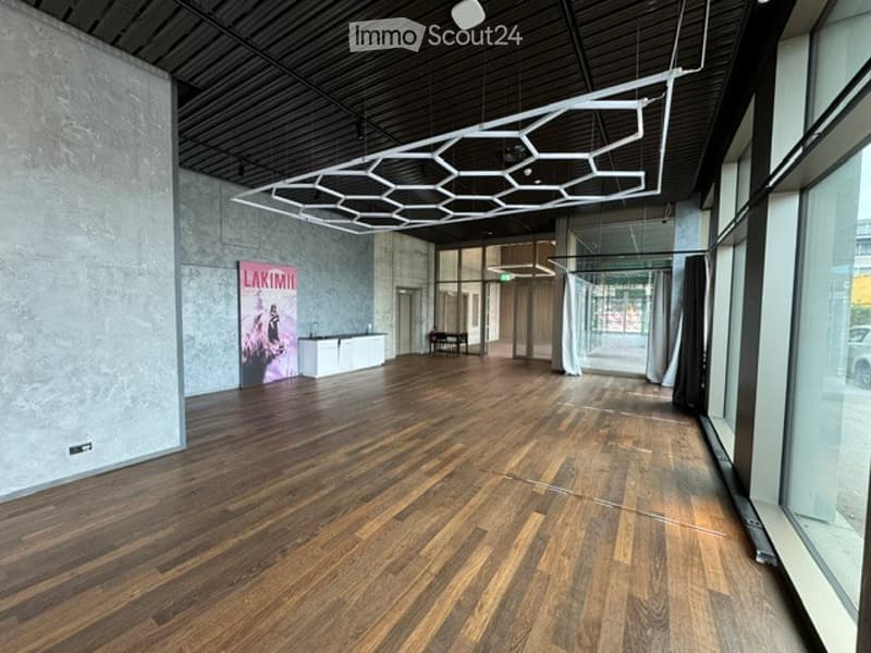 Empty room with wooden flooring, large windows, and a modern geometric light fixture on the ceiling. Minimalist design with a reception desk and a small seating area visible.