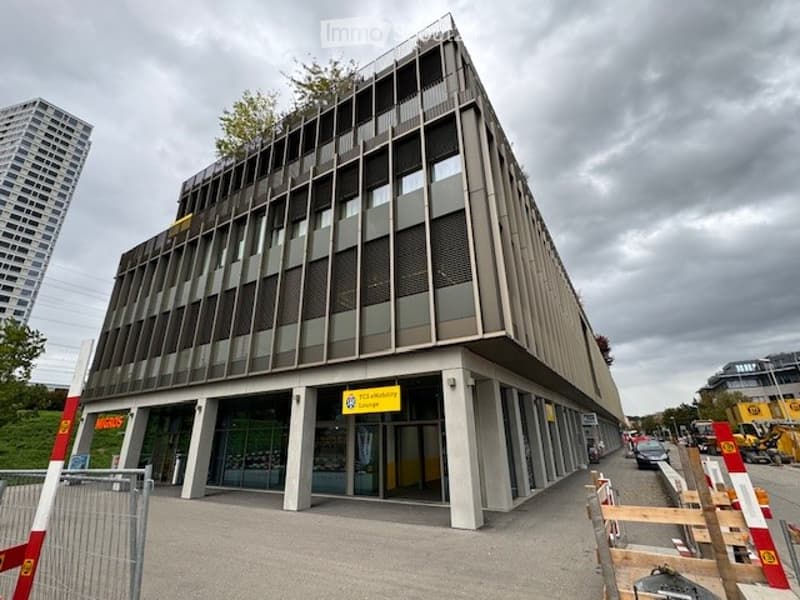 Modern multi-story building with a sleek, angular design and large glass windows. The entrance features a sign and a paved area with barriers.