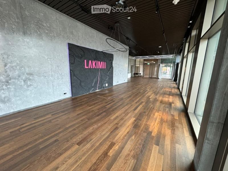 Empty hallway with polished wooden flooring, exposed concrete walls, and modern lighting fixtures. A large sign reading "LAKIMI" is mounted on the wall.