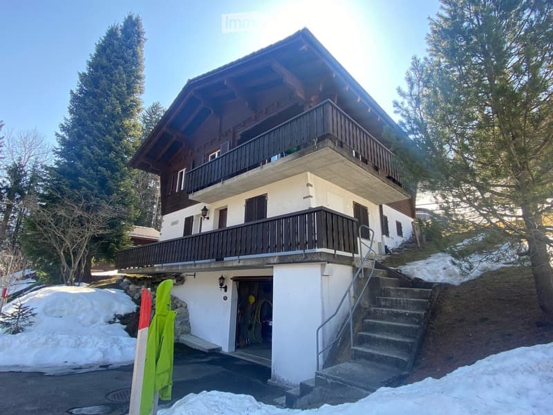 Two-story house with a wooden balcony, white exterior, and a small garage entrance. Surrounded by trees and snow-covered ground.