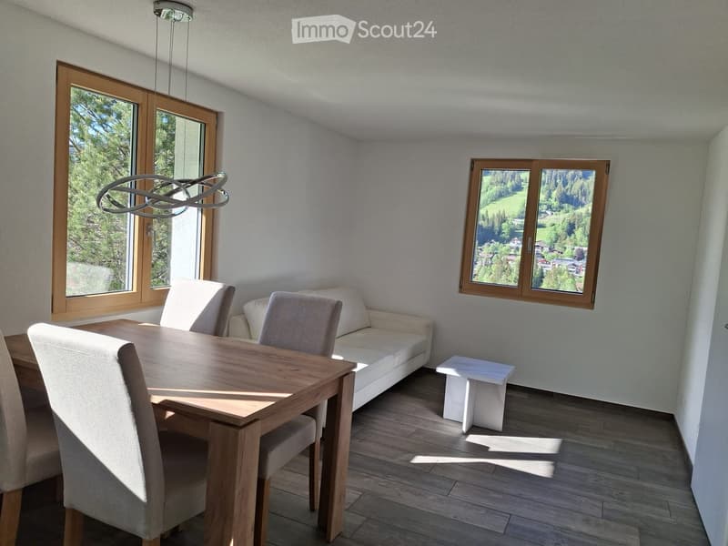Bright living and dining area with large windows, a sofa, a dining table with chairs, and a small white stool.