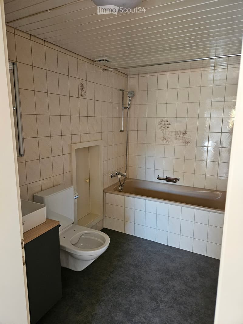 Modern bathroom with beige tiled walls, a bathtub, toilet, sink, and showerhead. Features a dark grey carpeted floor and a wooden vanity.