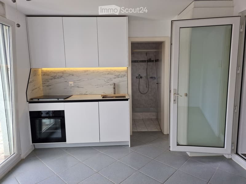 Modern kitchen with white cabinets, marble countertop, built-in oven, and a small shower area visible through an open doorway.